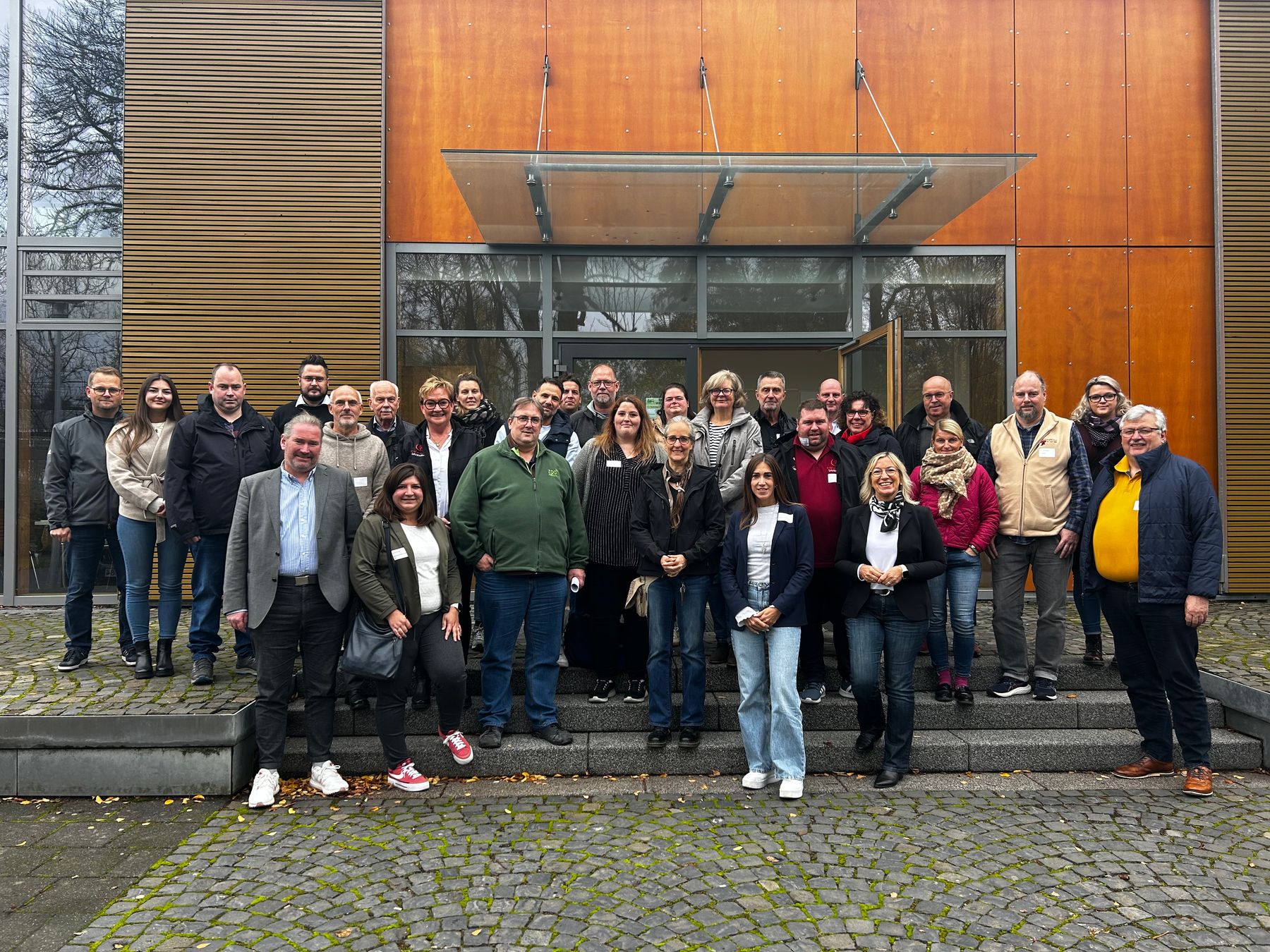 A group of about 30 adults stand together on stone steps outside a modern building with wooden panels and large glass doors, posing and smiling for a group photo.